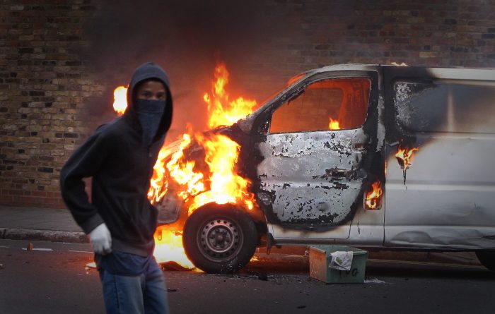 LONDON, ENGLAND - AUGUST 08: A hooded youth walks past a burning vehicle in Hackney on August 8, 2011 in London, England. Pockets of rioting and looting continues to take place in various boroughs of London this evening, as well as in Birmingham, prompted by the initial rioting in Tottenham and then in Brixton on Sunday night. It has been announced that the Prime Minister David Cameron and his family are due to return home from their summer holiday in Tuscany, Italy to respond to the rioting. Disturbances broke out late on Saturday night in Tottenham and the surrounding area after the killing of Mark Duggan, 29 and a father-of-four, by armed police in an attempted arrest on August 4. (Photo by Peter Macdiarmid/Getty Images)
